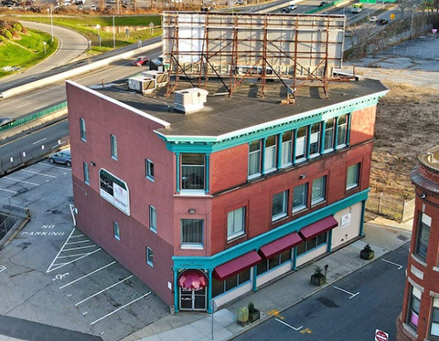 Three-story brick building with teal trim, red awnings, and a large billboard on the roof. Nearby parking area.