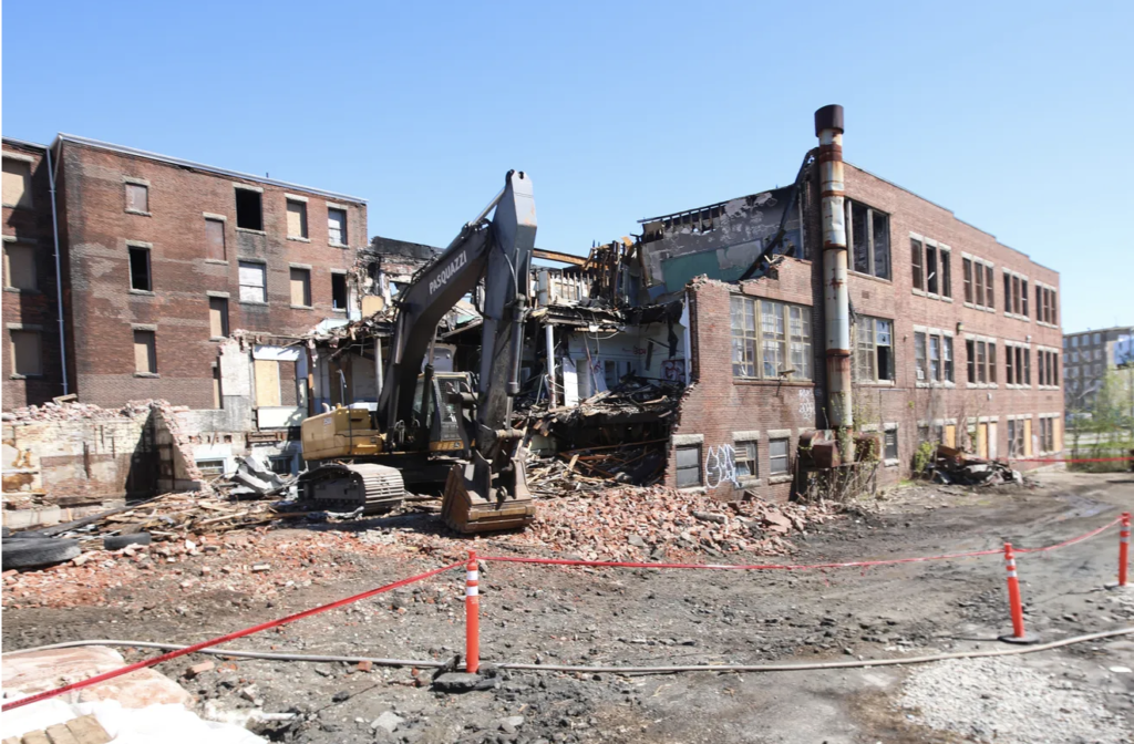 Excavator demolishes a partially collapsed brick building, surrounded by debris and construction barriers.