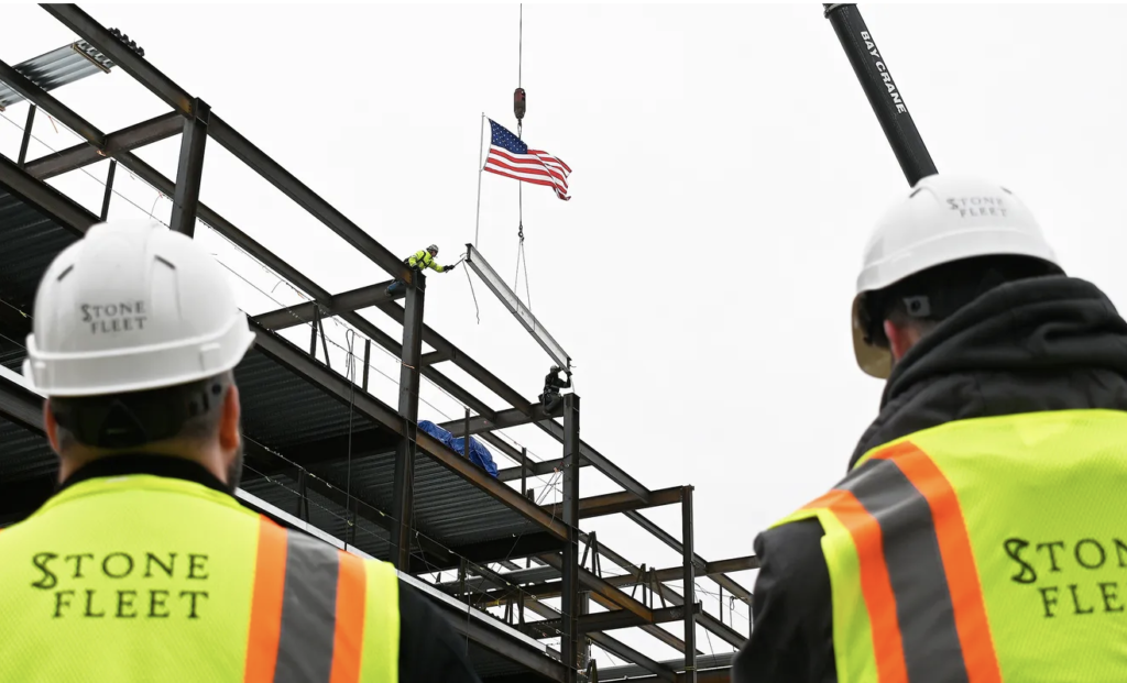 Construction workers watch as a crane lifts a beam with an American flag attached to it.