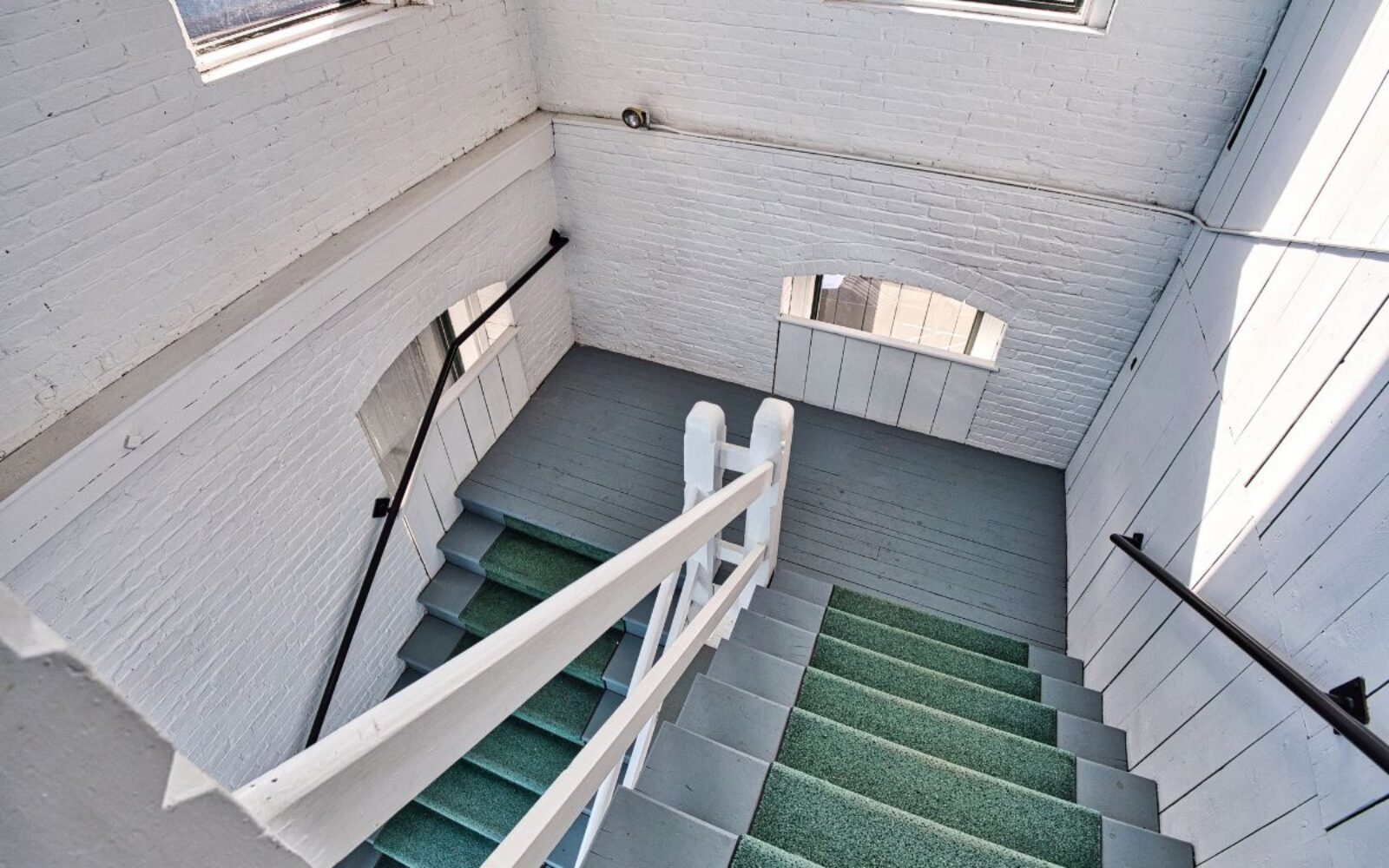 Staircase with green carpet, white railing, and brick walls, leading to a well-lit landing.