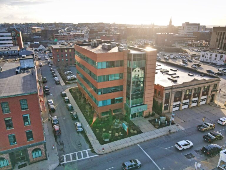 Aerial view of a modern office building surrounded by parked cars and cityscape in the background.