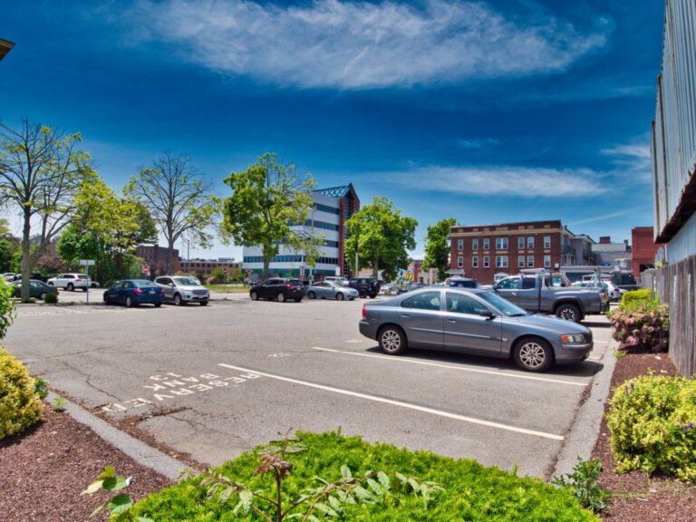 Parking lot with several cars, trees, and buildings under a clear blue sky.
